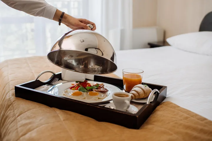 Plateau de petit-déjeuner servi sur un lit, avec café, jus d’orange, viennoiseries et œufs sous une cloche argentée
