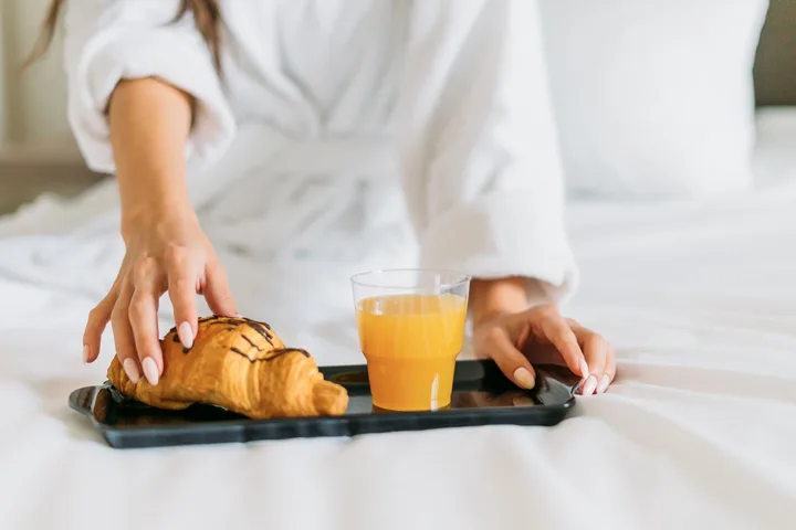 Deux personnes en peignoir blanc posent un plateau avec un croissant et un verre de jus d’orange sur un lit